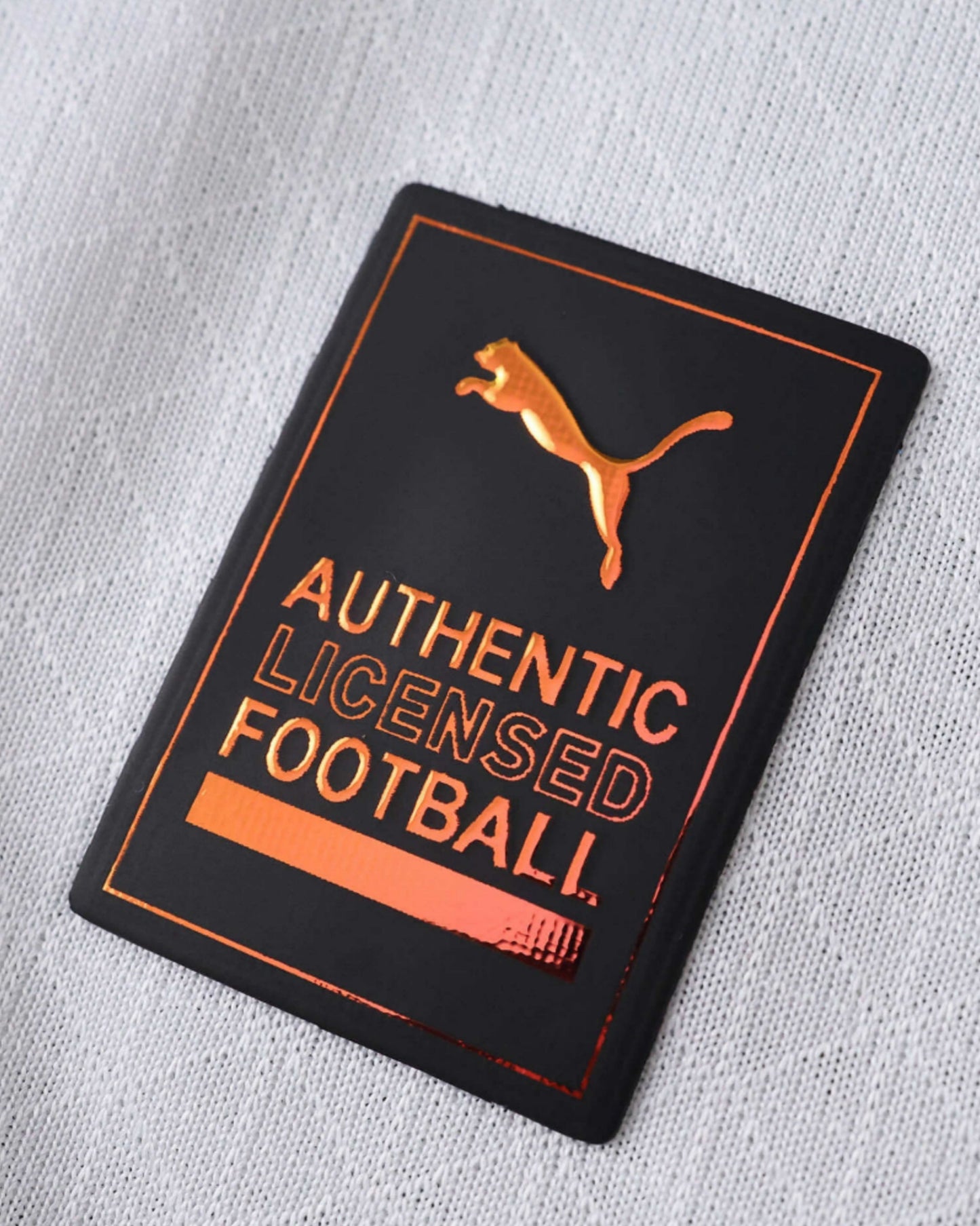 Close-up of a black and copper Puma authentic licensed football patch on a white textured jersey.