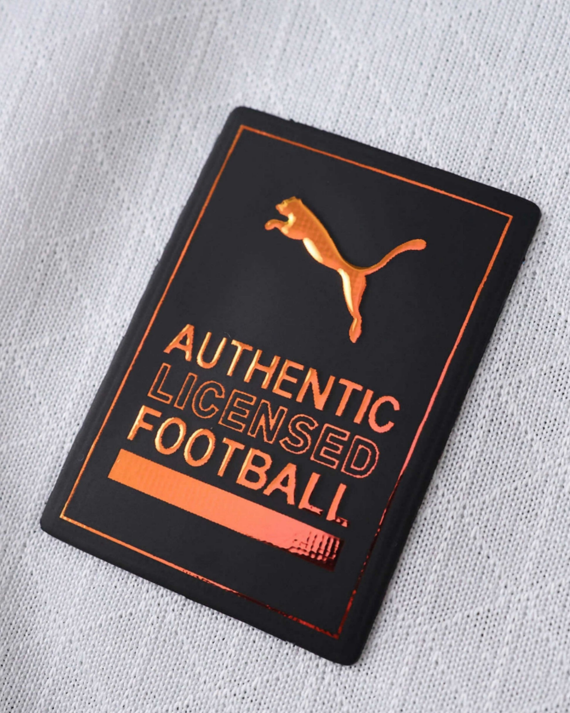 Close-up of a black and copper Puma authentic licensed football patch on a white textured jersey.